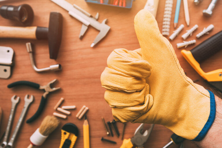 Gloved hand giving a thumbs-up with various tools on a wooden surface.