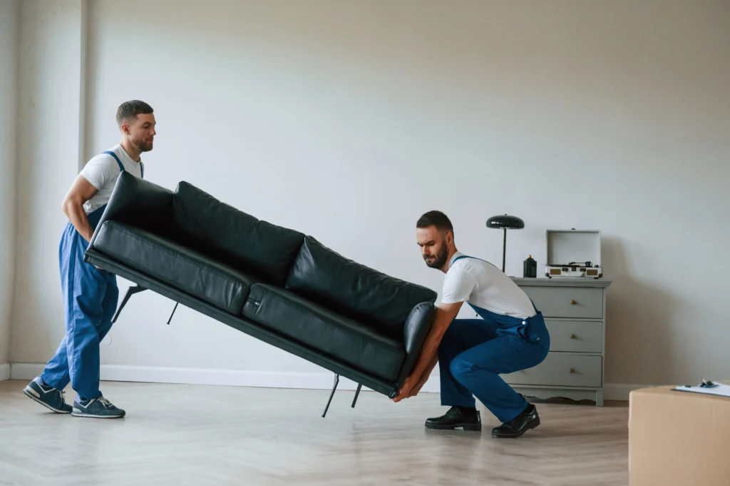 Zwei Umzugshelfer heben ein schwarzes Sofa in einer leeren Wohnung.